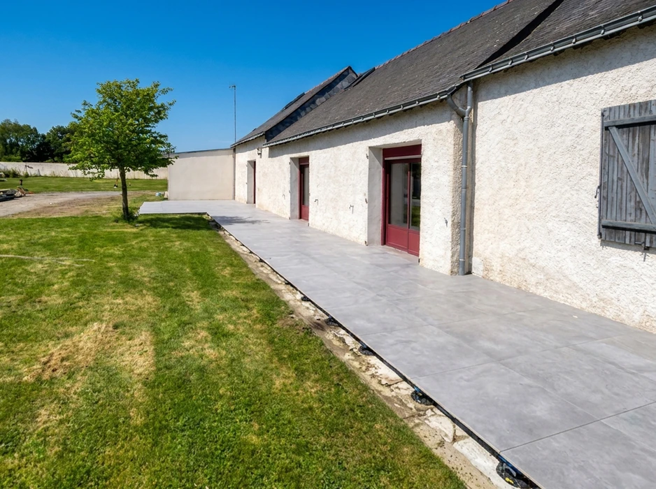 Terrasse extérieure en dalles grès cérame gris clair, posées en pose sur plots ou lit de sable, longeant la façade d'une longère rénovée en crépi blanc